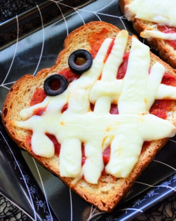 Black spider web tray with a slice of toast with tomato sauce and melted piece of cheese to look like a mummy with olive eye balls.