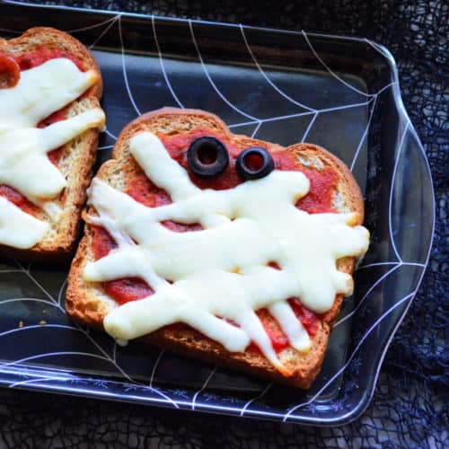 marinara, mozzarella, and black olives topping toast to look like mummy face in spider web dish.