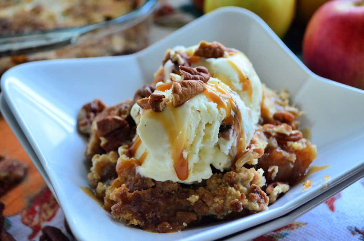 square dish of crumbled looking light brown cake with pecans topped with ice cream and caramel.