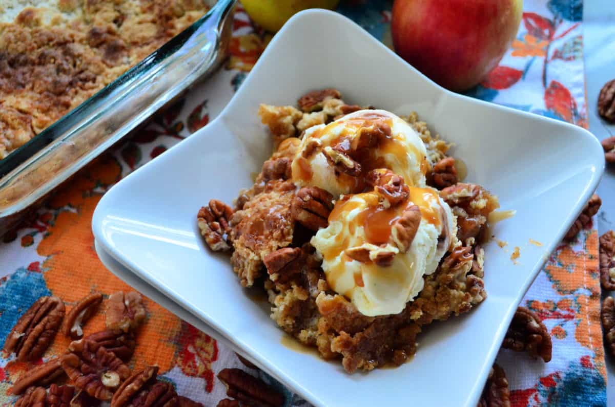 square dish of crumbled looking light brown cake with pecans topped with ice cream and caramel.