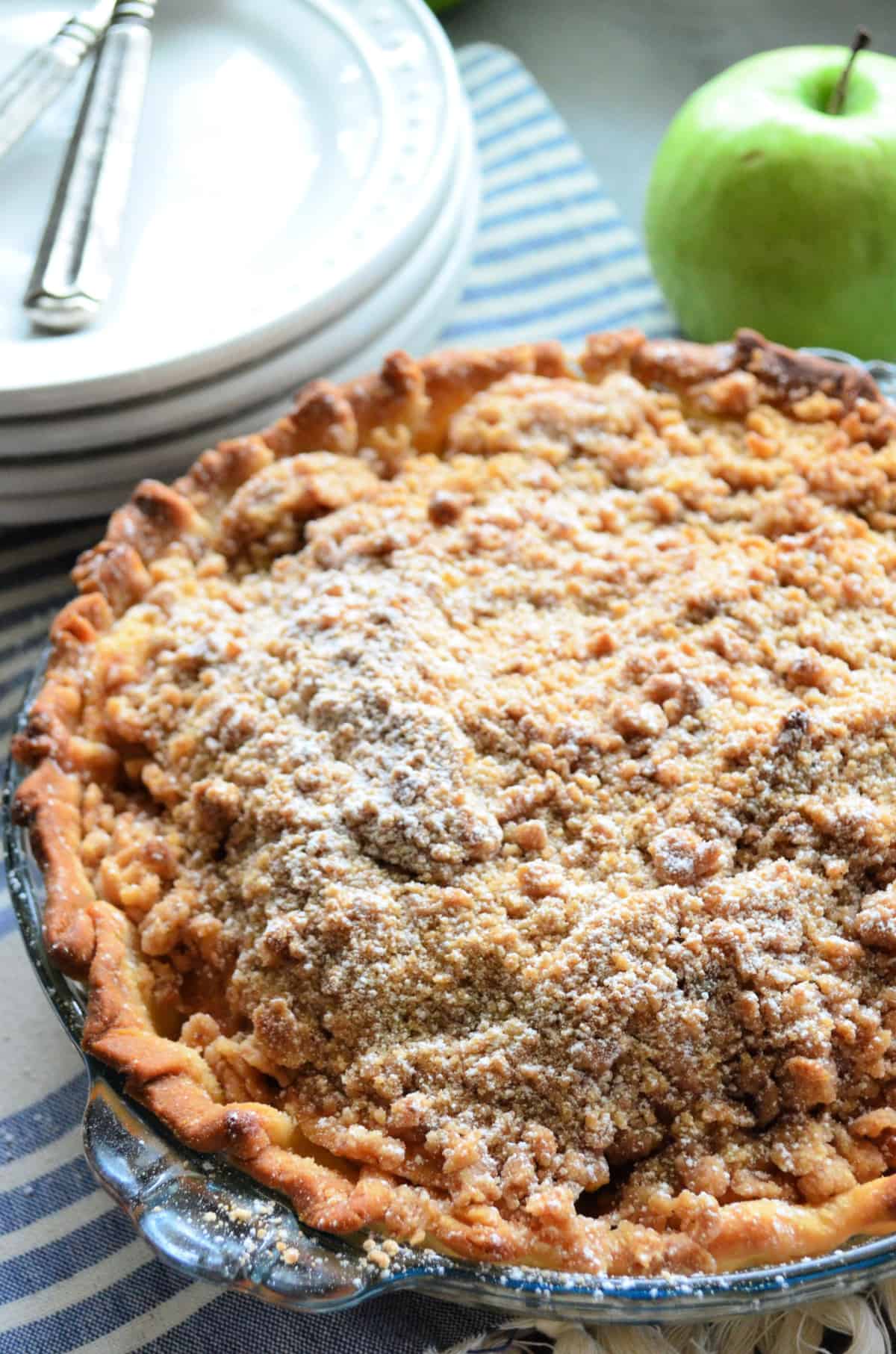 Pie with golden-brown crust and crumble topping dusted in powdered-sugar in front of a green apple.