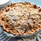 Pie with golden-brown crust and crumble topping dusted in powdered-sugar on tablecloth.