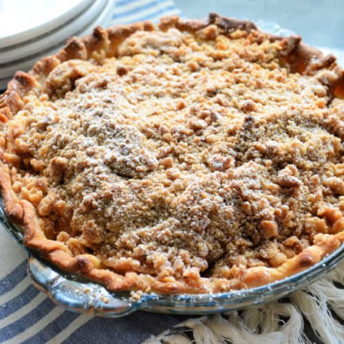 Pie with golden-brown crust and crumble topping dusted in powdered-sugar on tablecloth.