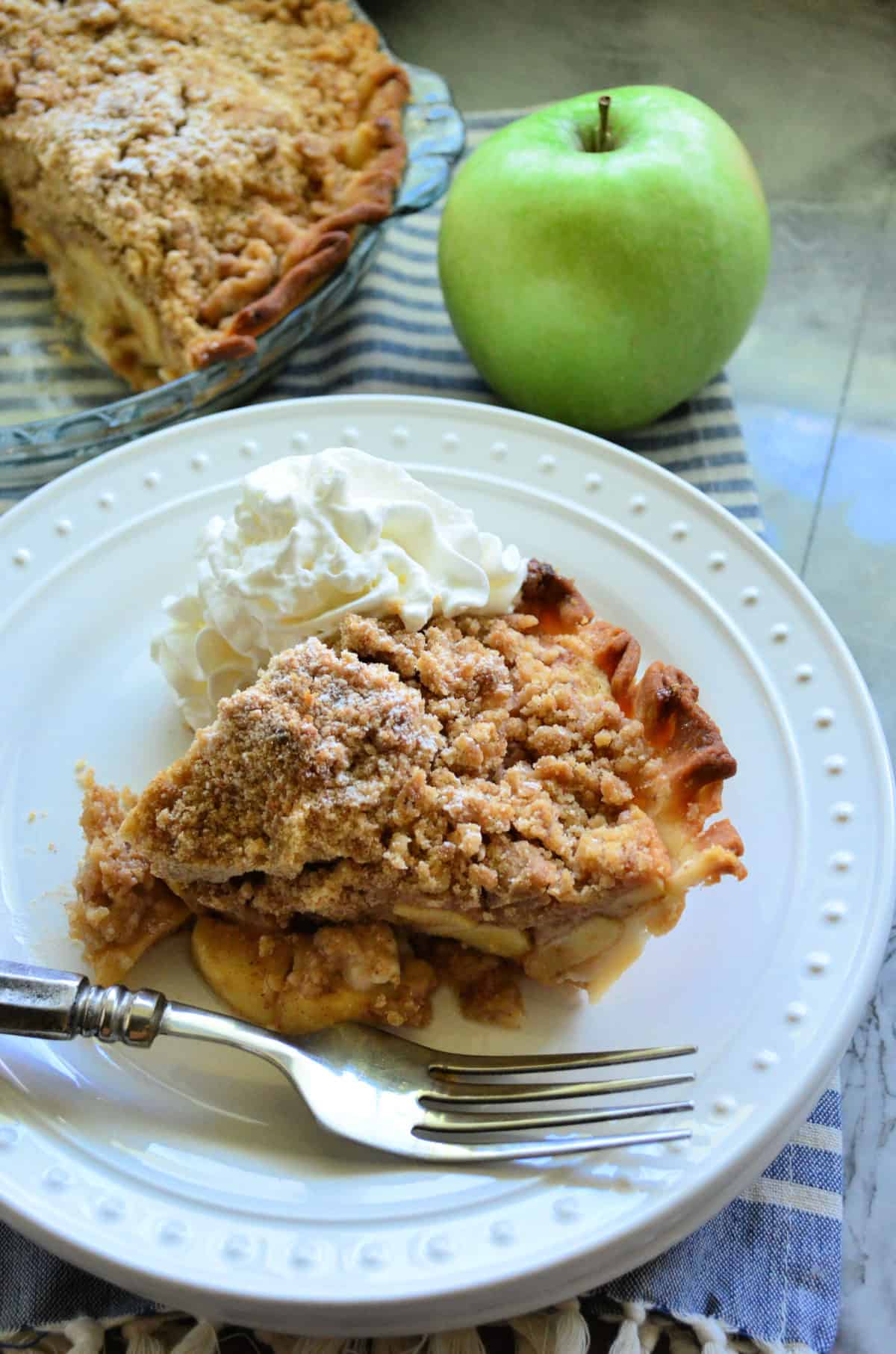 Plated slice of apple pie with fork and whipped cream next to fresh green apple.