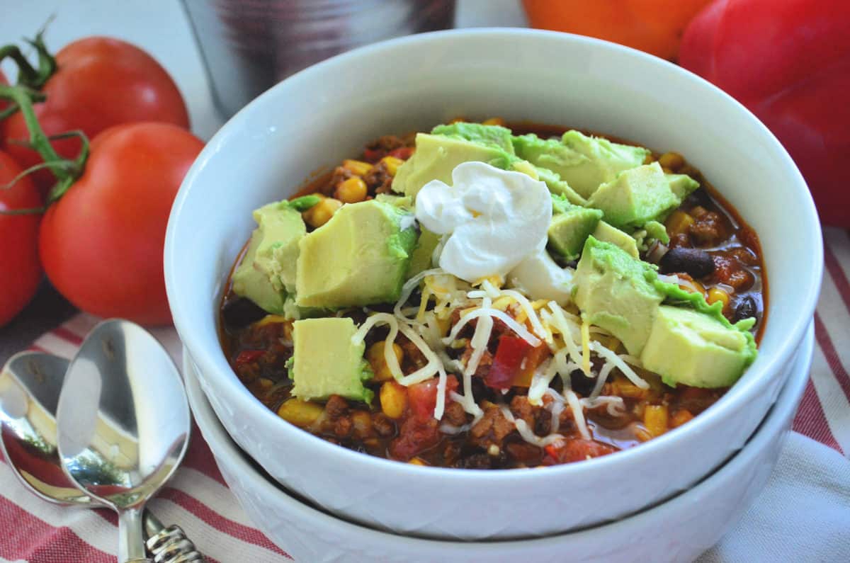 bowl of chili topped with avocado and shredded cheese on tablecloth with spoons and tomatoes.