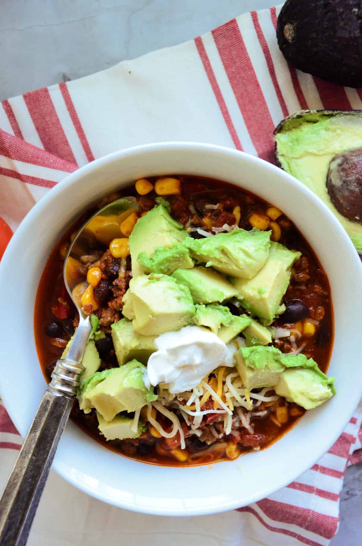 bowl of chili with spoon topped with avocado and shredded cheese on tablecloth with avocado half.