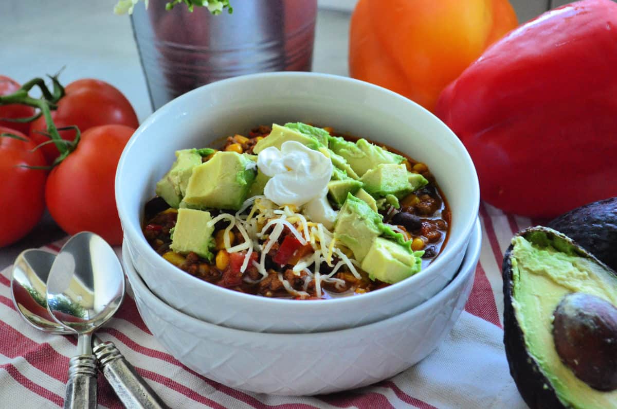 chili bowl topped with avocado and shredded cheese on tablecloth with avocado, tomato, and bell peppers.