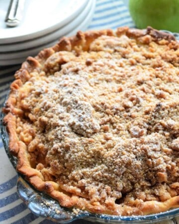 Pie with golden-brown crust and crumble topping dusted in powdered-sugar in front of a green apple.