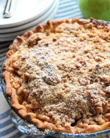 Pie with golden-brown crust and crumble topping dusted in powdered-sugar in front of a green apple.