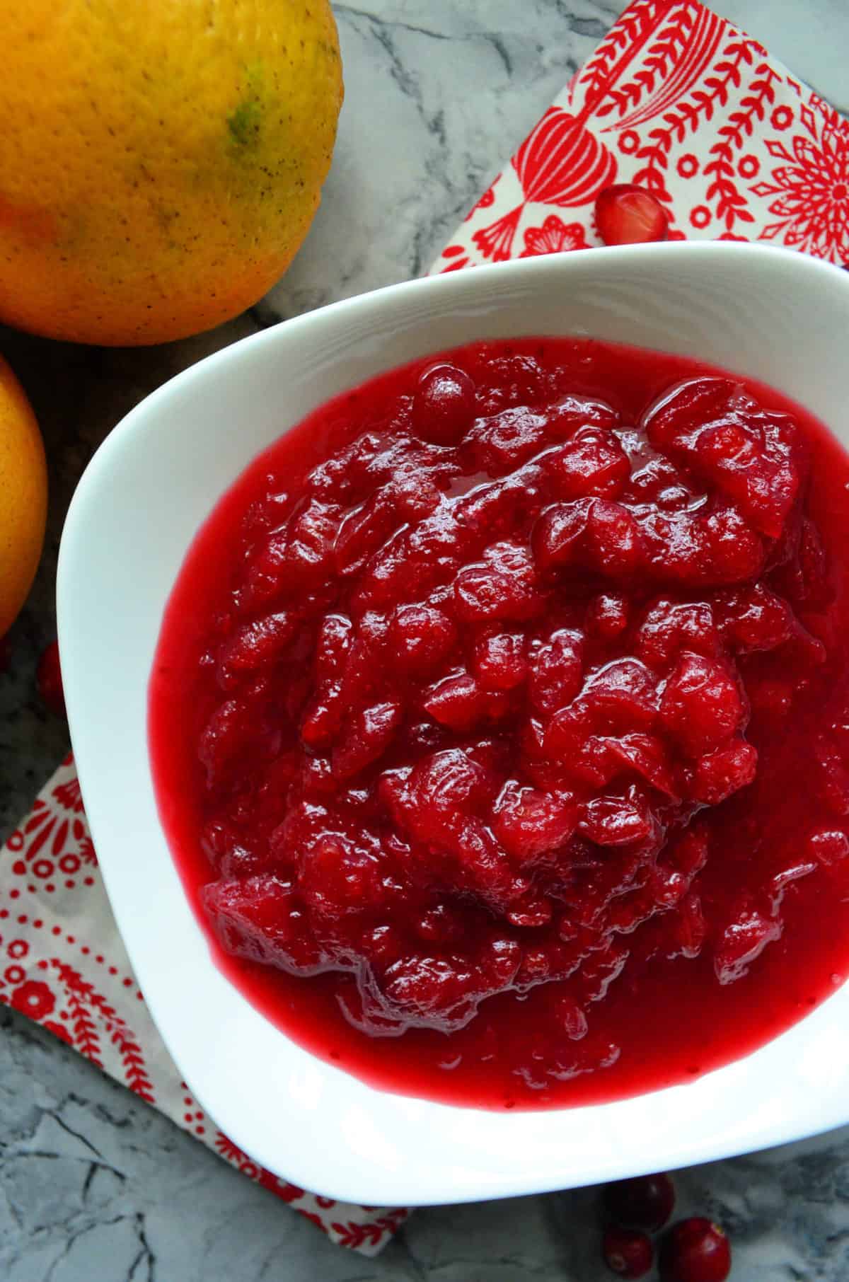 Top view of part of bowl of cranberry sauce on red and white tablecloth next to fresh oranges.