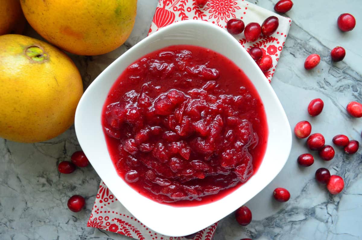 top view of chunky red cranberry sauce in white bowl on countertop with cranberries and fresh oranges.