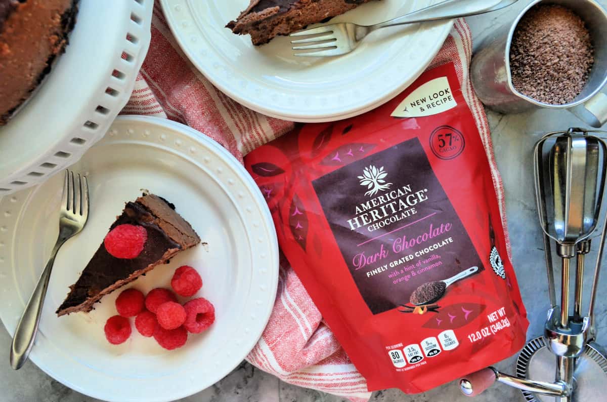 American Heritage Chocolate package next to plated cheesecake with raspberries, cup of brown powder, and whisk.