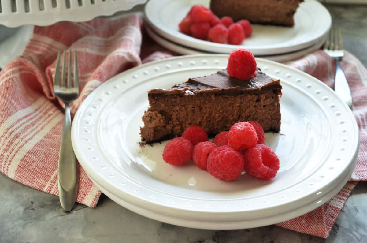 Plated slice of dark chocolate cheesecake with fresh raspberries on red tablecloth with forks.