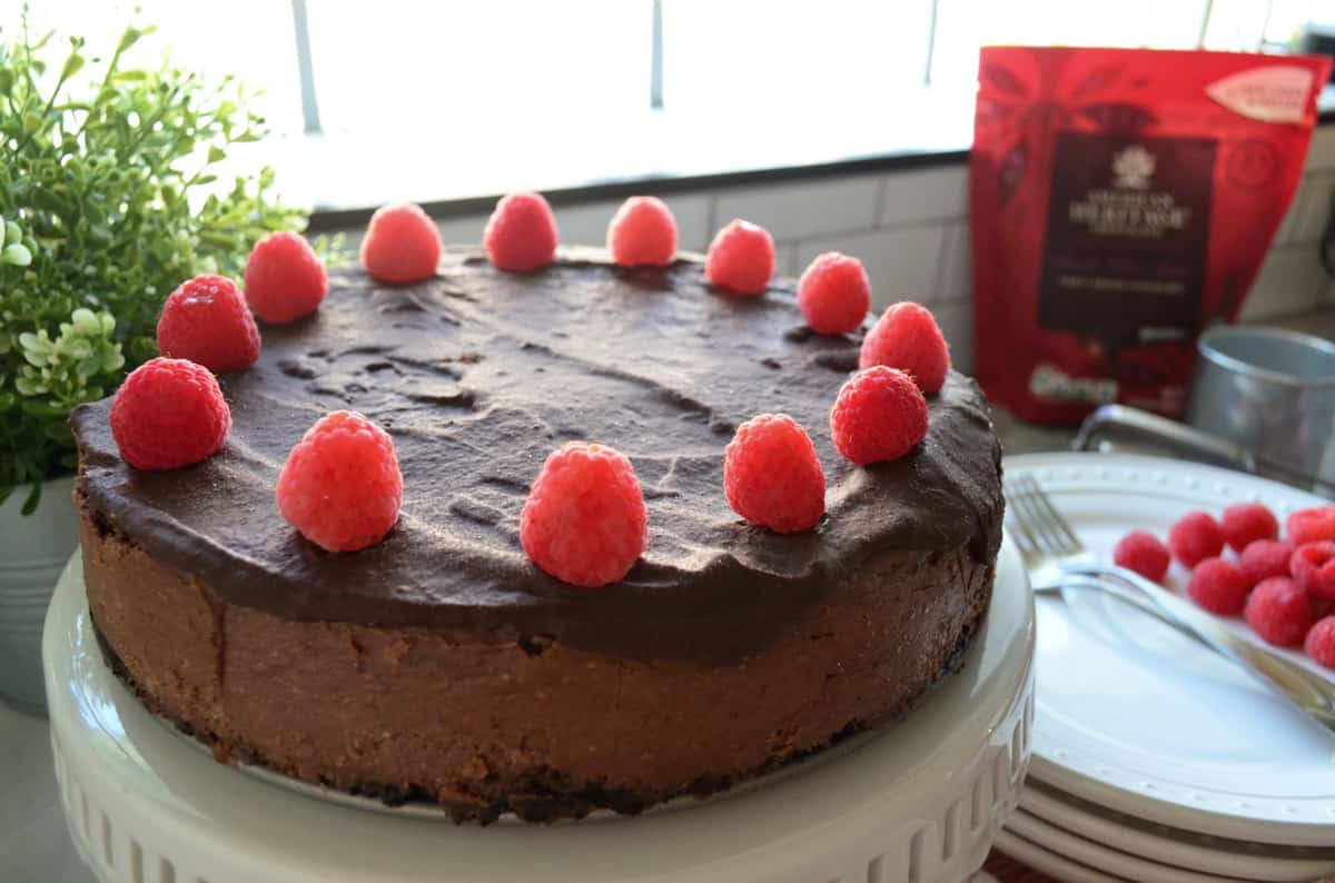 Dark Chocolate Cheesecake decorated with circle of raspberries on cake stand in front of window.