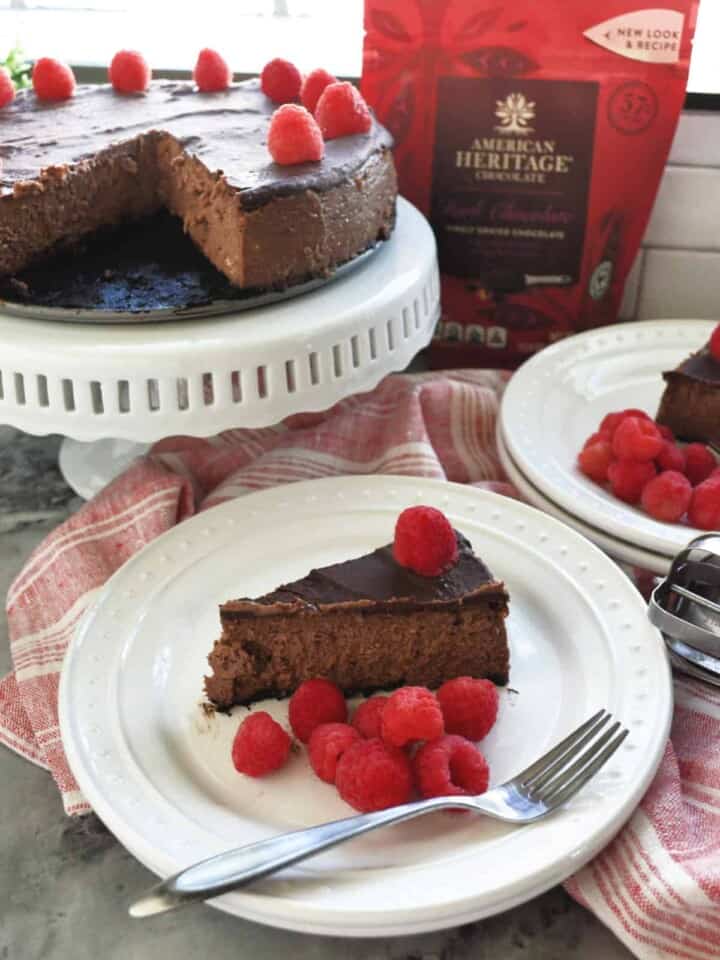 Slice of plated Dark Chocolate Raspberry Cheesecake in front of remaining cheesecake and bag of chocolate.