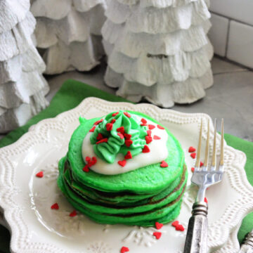 Green pancakes on white snowflake plate topped with green and white whipped cream with red sprinkles.