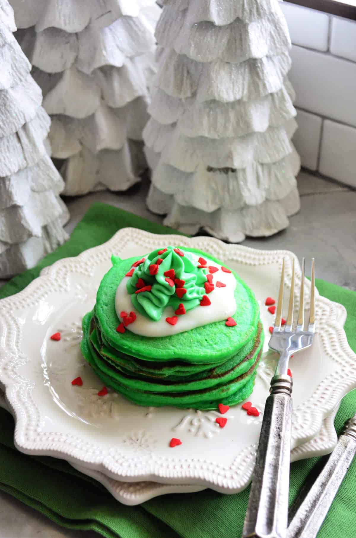 stack of grinch pancakes on white decorative plate next to fork in front of white tree table decor.