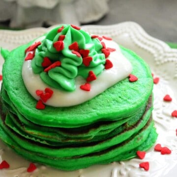 Green pancakes on white snowflake plate topped with green and white whipped cream with red sprinkles.