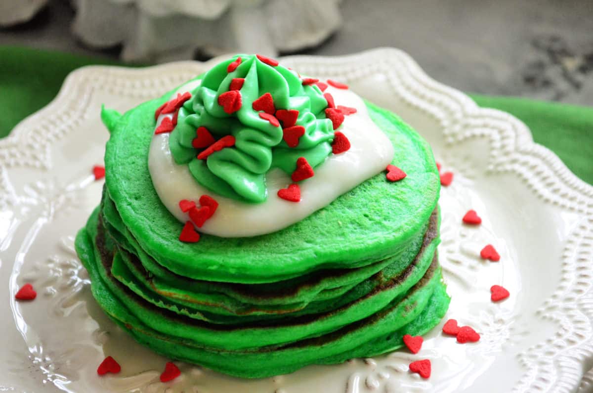 green pancakes with white icing, green whipped cream, and red heart sprinkles on white decorative plate.