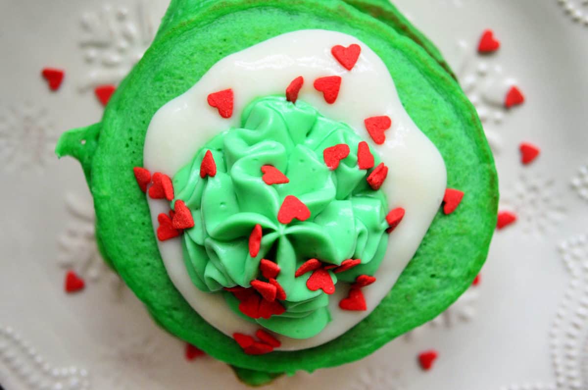 Top view of green pancakes with white icing, green whipped cream, and red heart sprinkles.