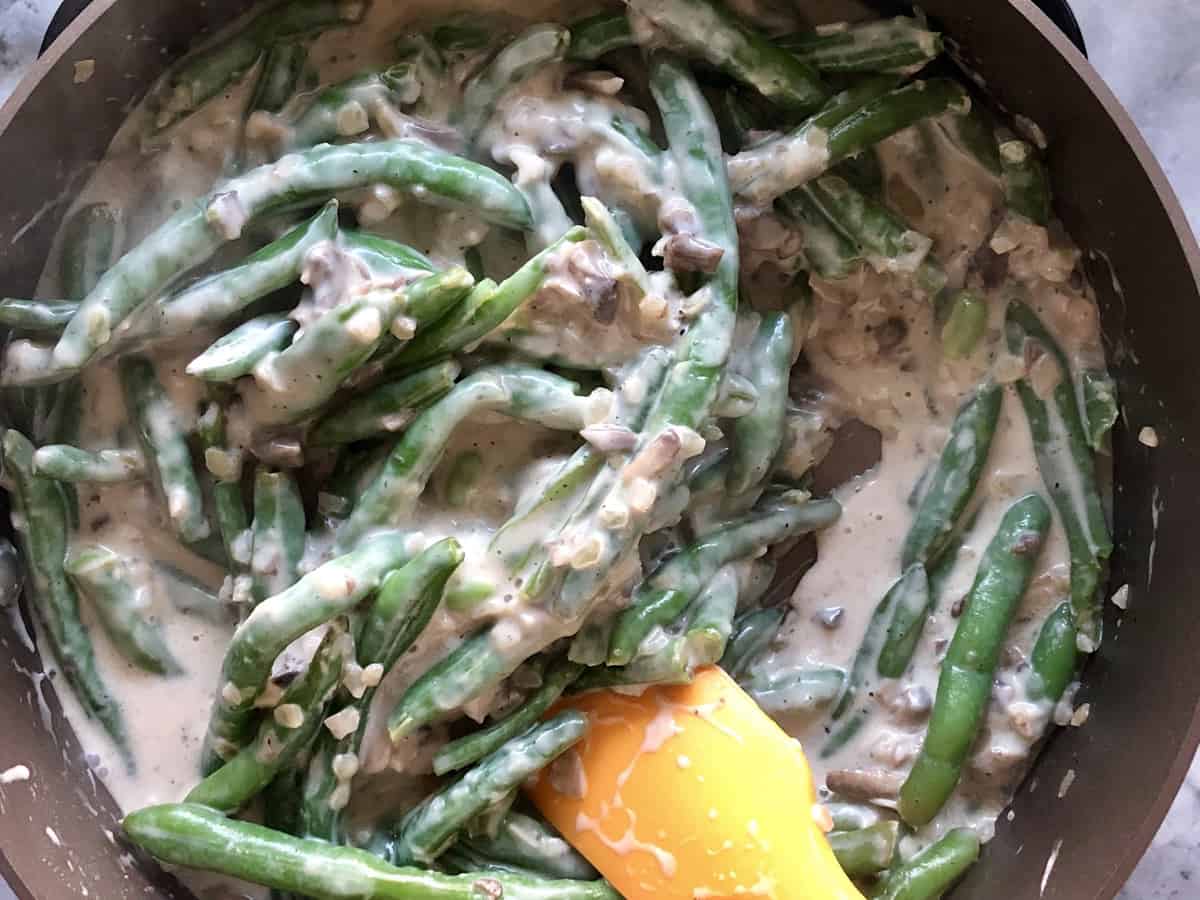 top view of green beans in pan being stirred with white sauce and chopped mushrooms with spatula.