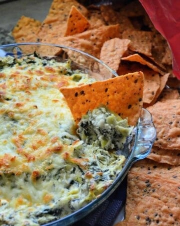 Bowl of Spinach Artichoke Dip with melted cheese on top and chip dipped into bowl.