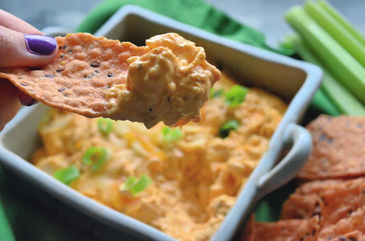 hand holding chip dipped in cheesy buffalo chicken dip with dip, celery, and chips in background.