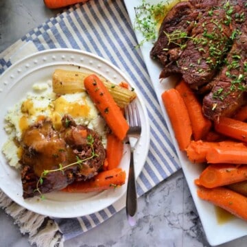 Plated Pot roast with mashed potatoes, celery, carrots, and gravy next to serving platter.