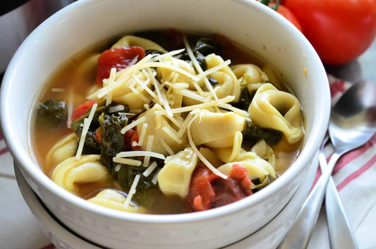 closeup of tortellini spinach and tomato soup topped with cheese in top bowl next to spoons.