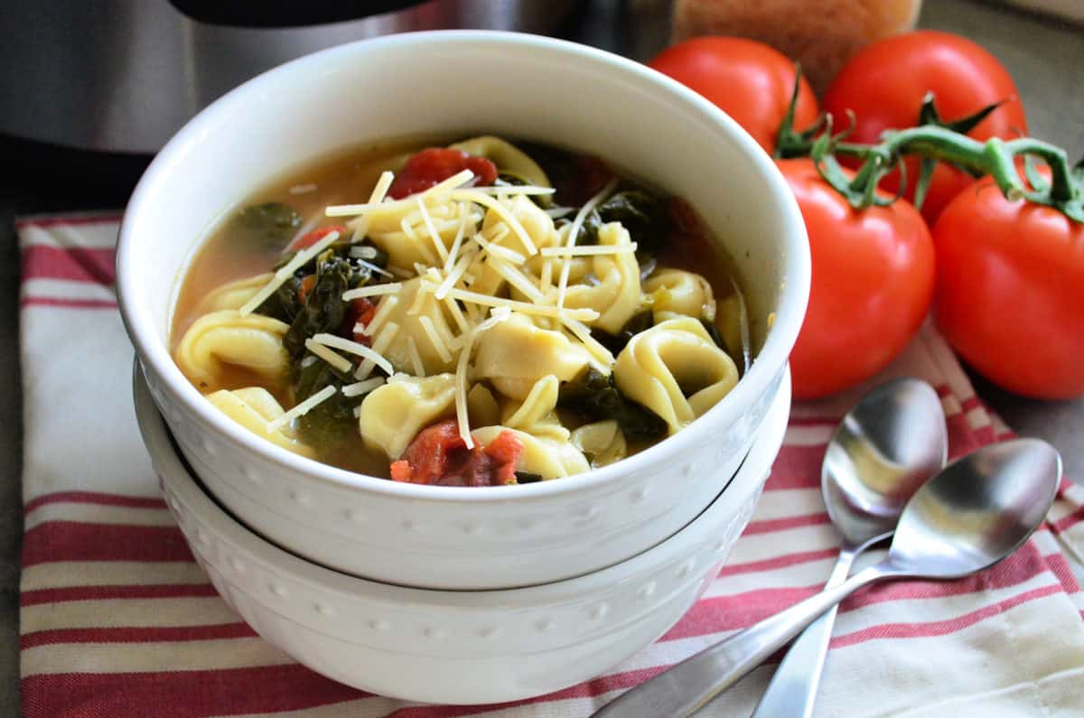 Two bowls stacked with tortellini spinach and tomato soup topped with cheese in top bowl next to spoons.