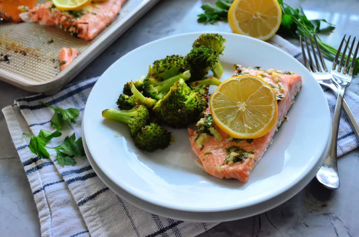 Plated cooked salmon topped with lemon wheel served with roasted broccoli next to forks.