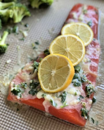 Large piece of raw salmon with three slices of lemon, butter, and herbs on a sheet pan.
