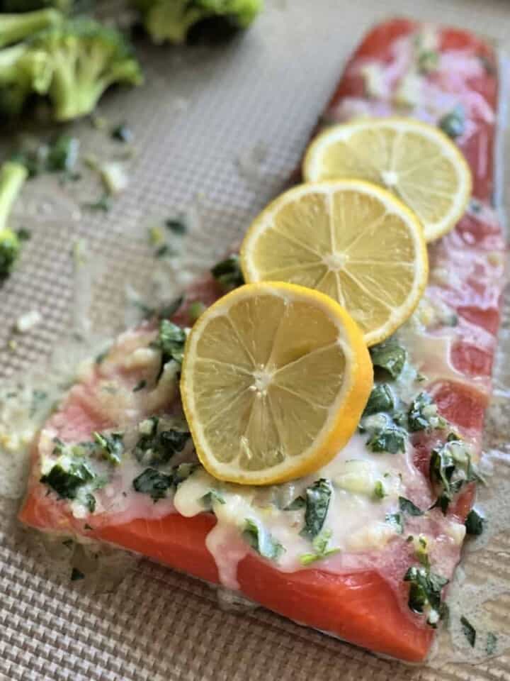 Large piece of raw salmon with three slices of lemon, butter, and herbs on a sheet pan.