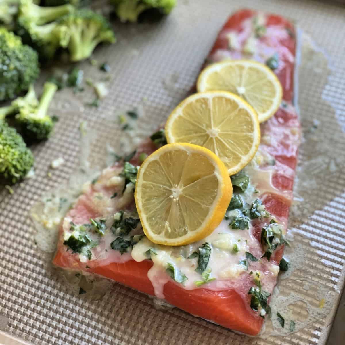 Large piece of raw salmon with three slices of lemon, butter, and herbs on a sheet pan.