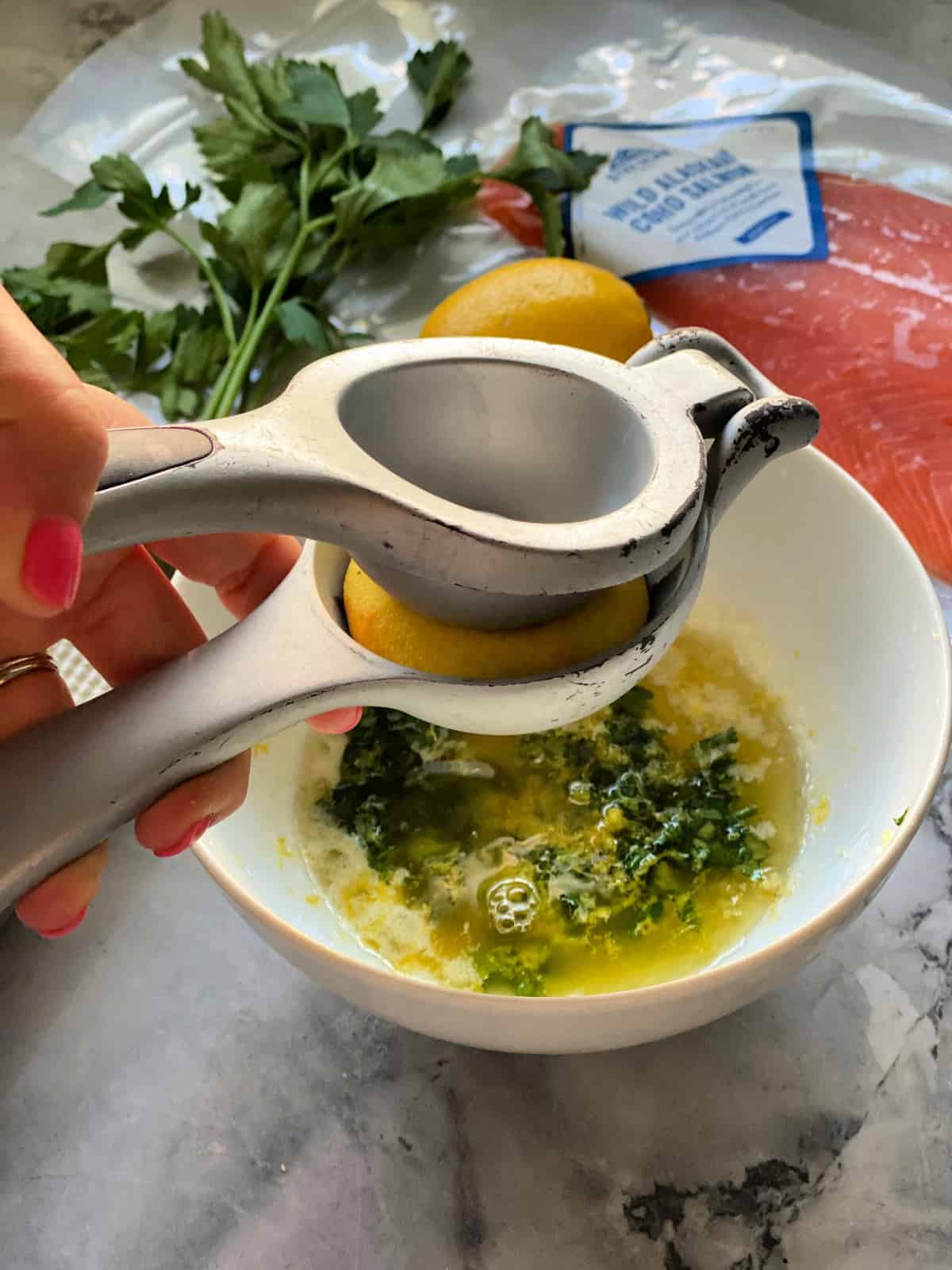 Hand using press to juice lemon into bowl with butter and parsley next to package of salmon.