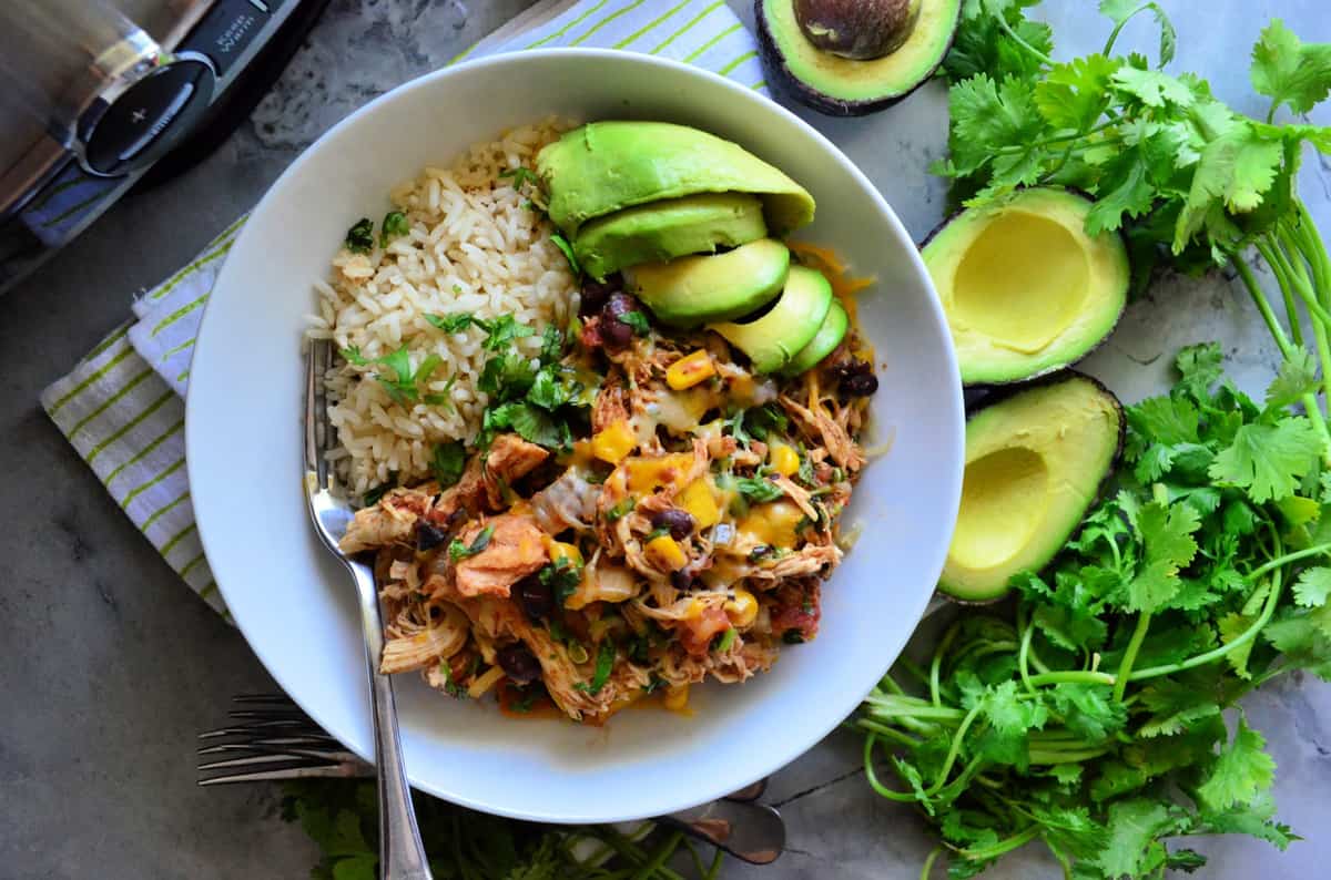 Top view of a white bowl filled with rice, shredded chicken, black beans, cheese, and avocados.