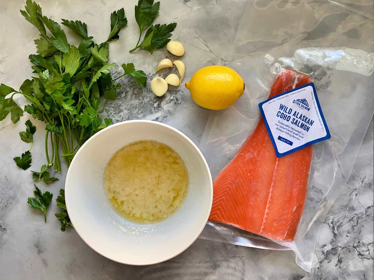 Top view of packaged salmon, melted butter, garlic cloves, parsley, and whole lemon on countertop.