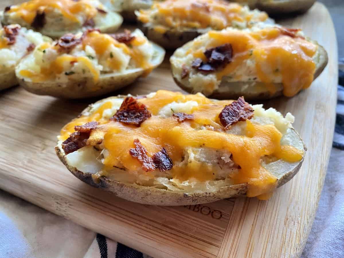 Side view of loaded potatoes with melted cheese and bacon on wood cutting board.