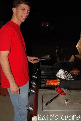 side view of man smiling and holding tongues as he cookes lobster tails and steak on the grill.