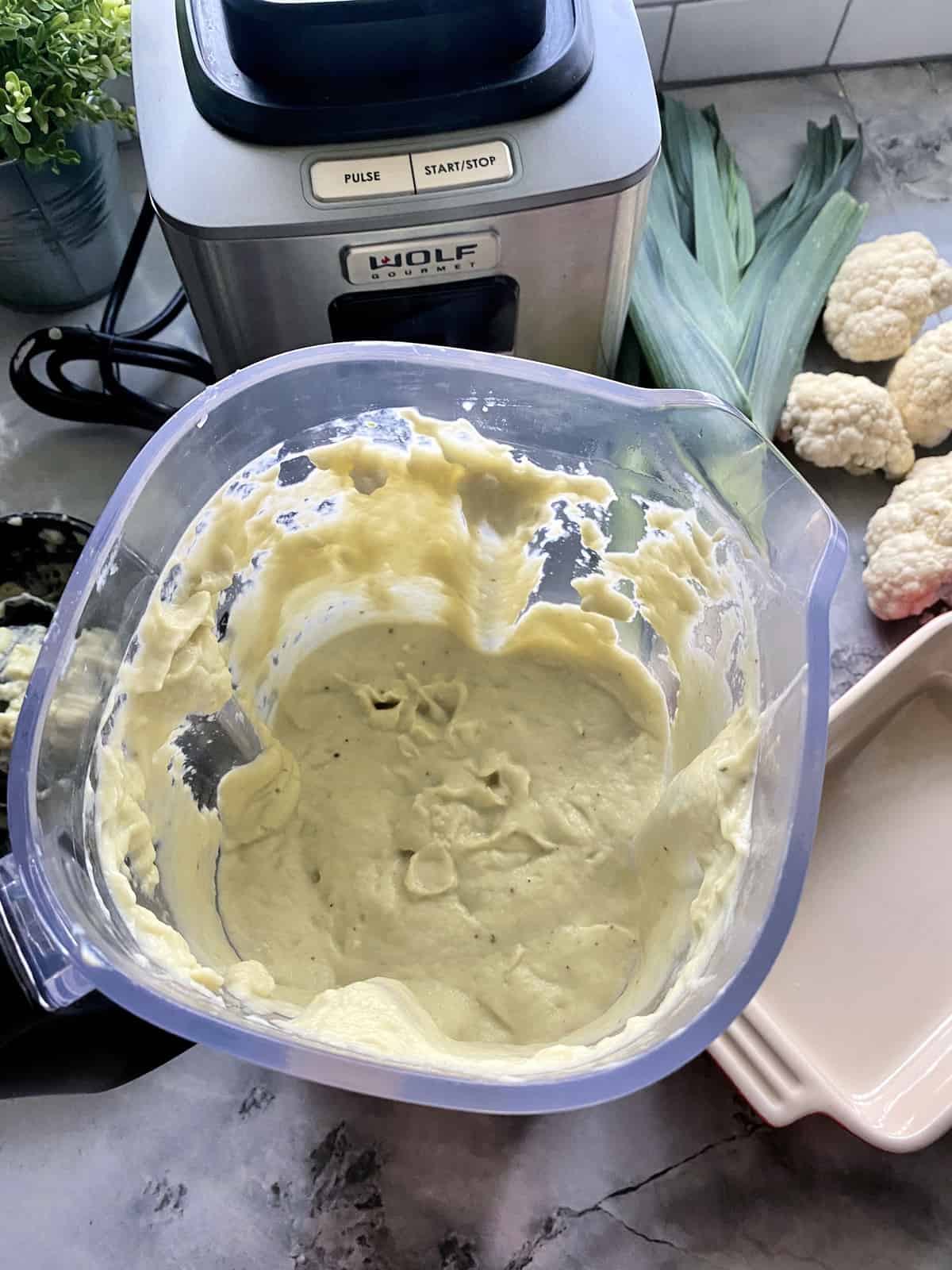 top view of Blended Cauliflower-Leek Puree inside blender next to cauliflower and leeks on counter.