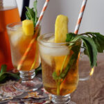two sunday glasses with golden colored tea garnished with mango slices, mind, and paper straws.