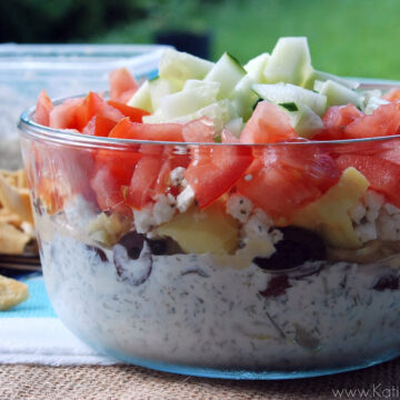 Side view of 7 layer greek dip in glass bowl topped with tomato and cucumber.