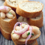 sliced mini french bread stacked and topped with white bean and red onion bruschetta and green herbs.