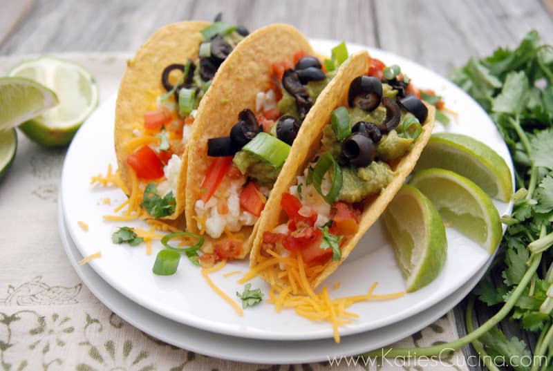 Three plated egg white tacos topped with olives, chives, tomatoes, and cheese garnished with lime and cilantro.