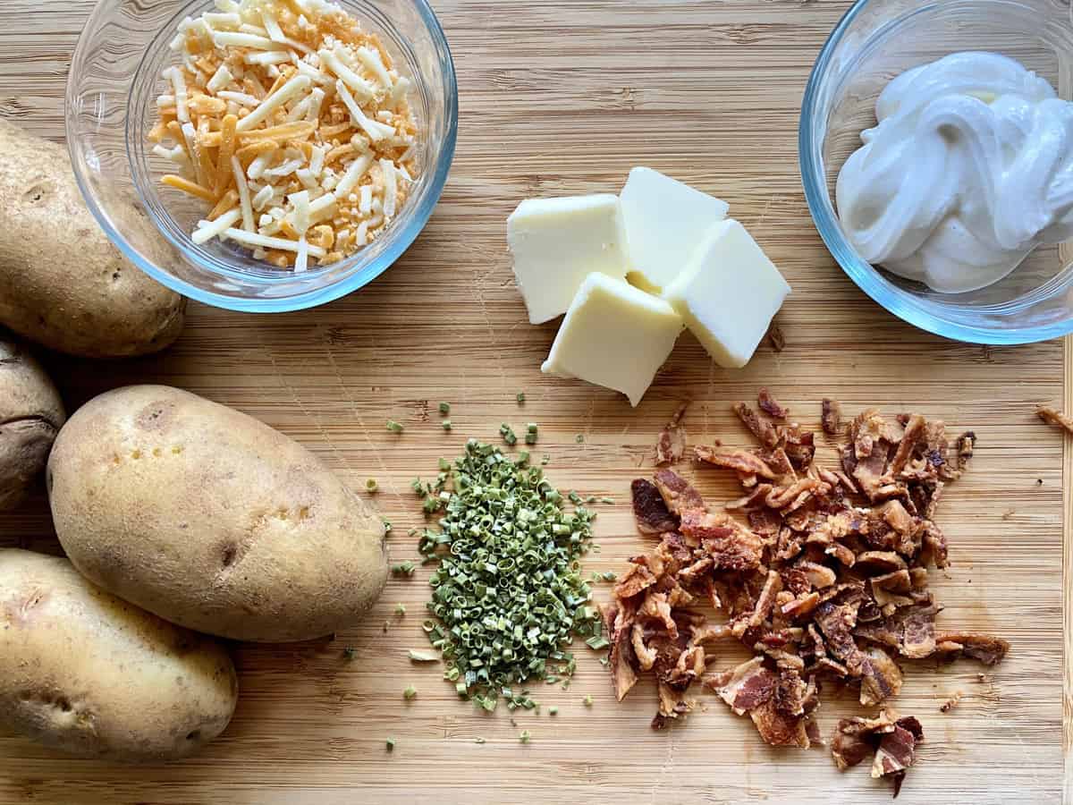 top view of bacon, sour cream, butter, chives, potatoes, and shredded cheddar on wood board.