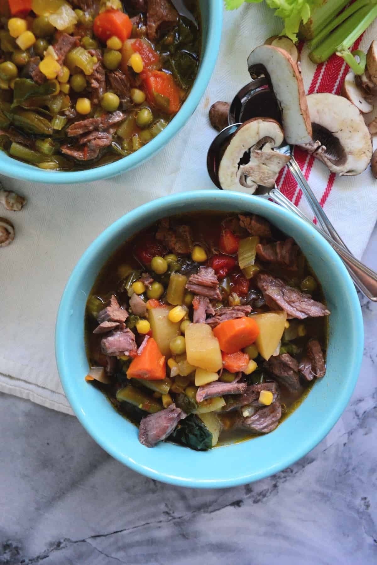 2 bowls of soup with beef, carrots, green beans, corn, and peas visible on countertop near mushrooms.