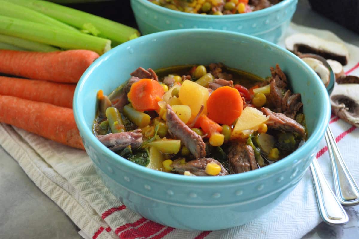 side view of soup with beef, carrots, green beans, corn, and peas visible on countertop with spoons.