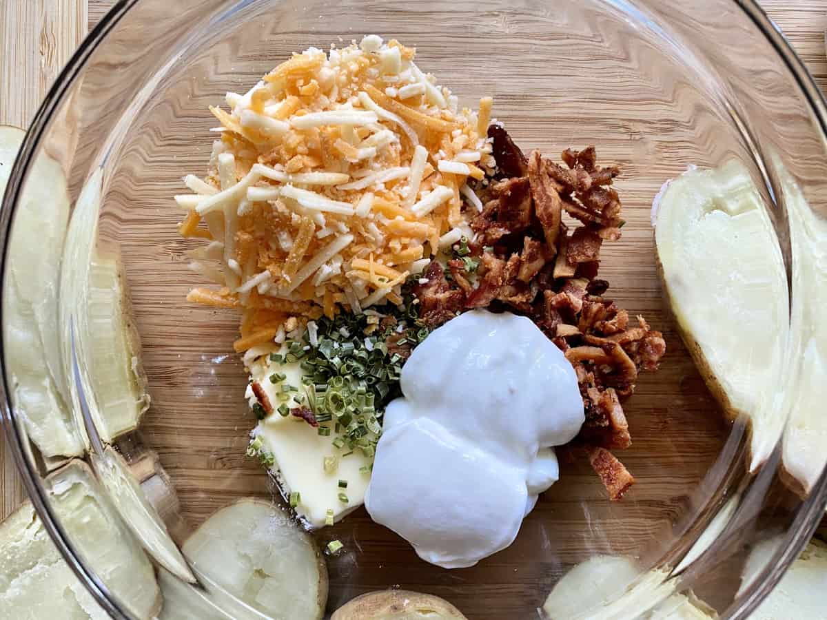 Top view of sour cream, butter, chives, bacon, and shredded cheddar in glass bowl next to halved potatoes.