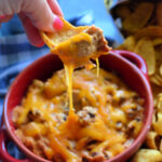 Manicured hand scooping up chili cheese dip from red bowl with frito lay chip.