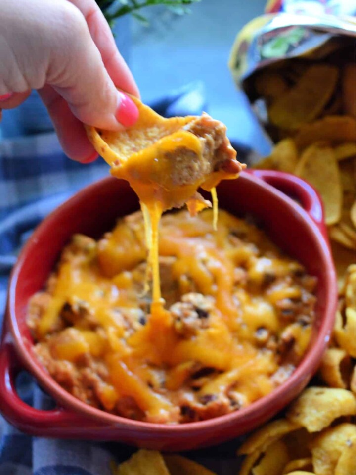 Manicured hand scooping up chili cheese dip from red bowl with frito lay chip.